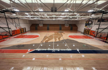 Indoor basketball court with a high-gloss finish on the wooden floor. The court has markings in red, white, and blue, with the key areas distinctly painted. There's a center circle with a logo , and basketball hoops along the sides of the court.
